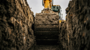 Yellow excavator with a dirt-covered shovel in a trench, viewed from the trench ground looking up at the overcast sky.