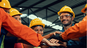 Several workers wearing orange jackets and yellow hardhats stack their hands together, representing teamwork.