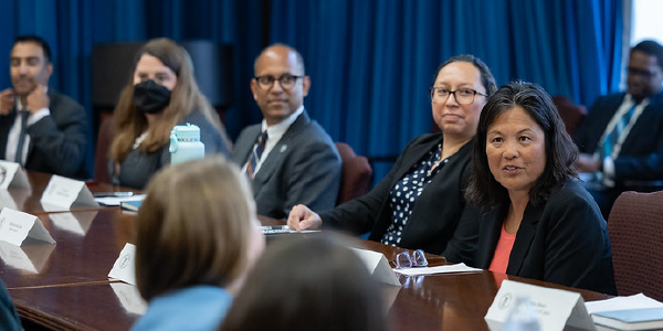 Acting Secretary Su listens to a woman seated across the table from her. They are surrounded by other men and women in professional clothes.