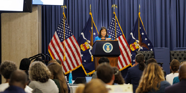 Acting Secretary Su speaks to a crowded room from a podium bearing the Department of Labor seal. She stands on a stage in front of U.S. and Labor Department flags. 