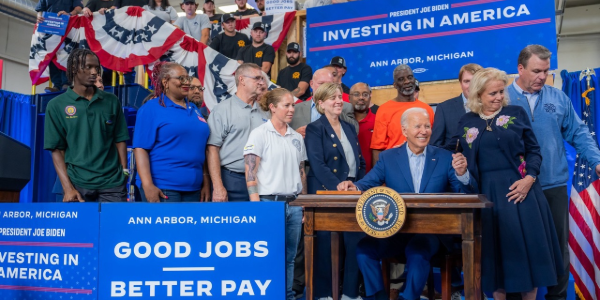 President Biden holds a pen after signing an executive order. He is surrounded by workers and officials. Signs read âInvesting in America â Good jobs, better pay.â