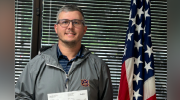 A smiling man holds a check while standing in front of an American flag.