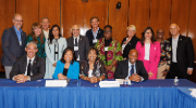 15 adults, all professionally dressed, gather for a group photo behind a conference table in a room with wood paneling. 