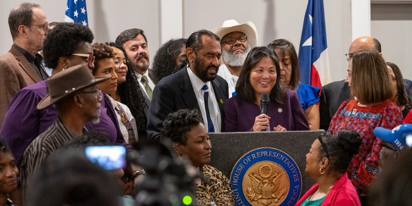 Acting Secretary Su speaks at a podium near Texas and US flags surrounded by workers of many racial and economic backgrounds.