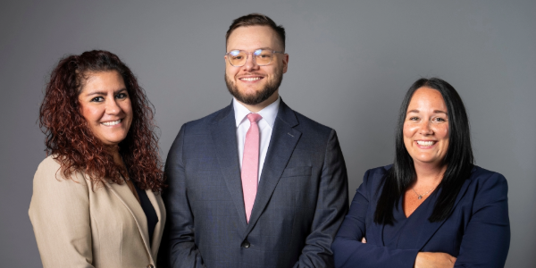 Three professionals standing together, with a plain gray background.