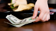 A person's hand removing a few dollars from a restaurant bill folder on a table.