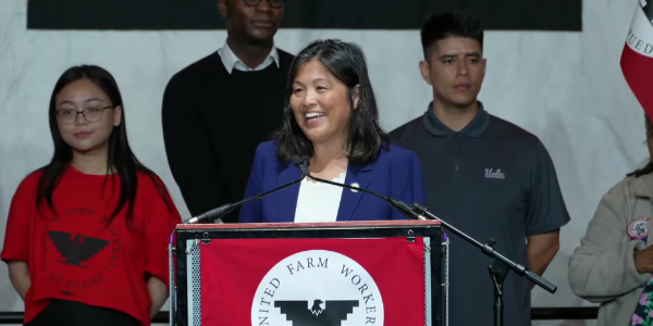 Acting Secretary Su speaks at a podium with the United Farm Workers logo. Behind her are several workers and young people.