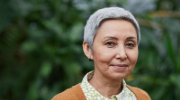 An older woman with fair skin and short, grey hair wearing a rust-colored sweater and green and white patterned button down.