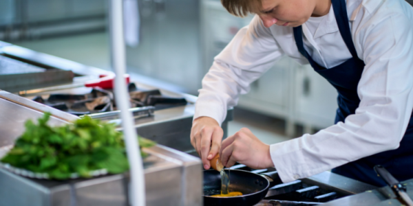 Young boy working in a restaurant kitchen cracking an egg into a frying pan.