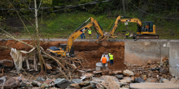 Workers in safety gear stand near a concrete slab in a sinkhole with concrete, trees and rocks beneath a road washed out by Hurricane Helene in Swannanoa, North Carolina. (Photo by Madeleine Cook, FEMA)