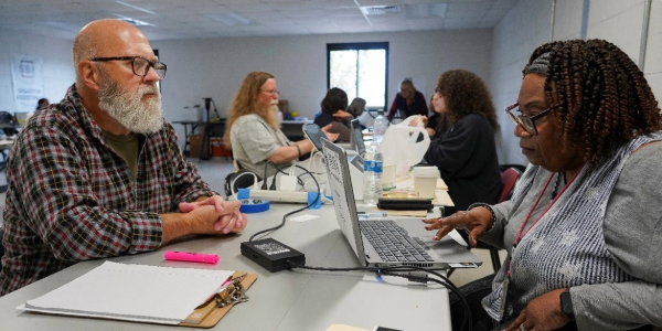 Bearded man with glasses in flannel shirt sitting across a table from a woman on a laptop as she assists the man with an application for financial assistance.