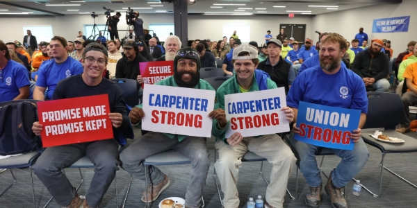 Four men seated in a crowd smile. They hold signs reading, “Promise made, promise kept,” “Carpenter strong,” and “Union strong.”