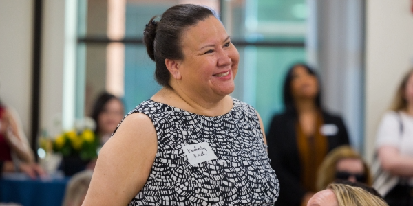 A woman wearing a nametag that says “Kimberly Rush” stands in a conference room, surrounded by other women. She is smiling.