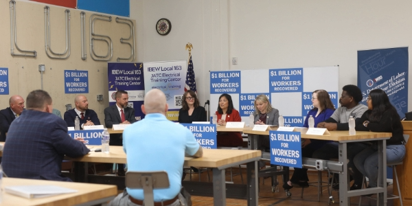 Acting Secretary Su and Wage and Hour Division Administrator Jessica Looman sit at a table with other adults. Signs around the table read “$1 billion for workers recovered.”