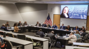 Young woman stands in large room filled with professionally dressed people seated at tables, and faces a projection screen that reads “Improving UI Customer Experience & Access”