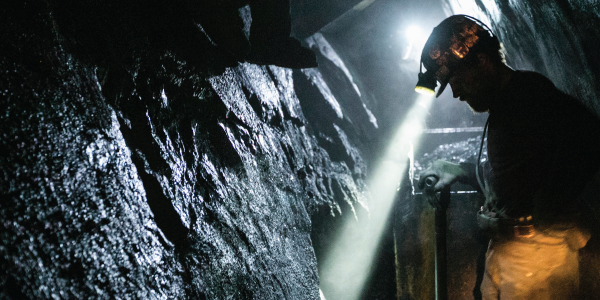 Coal miner in a dark coal mine with a lighted helmet looking down at the mine floor. 