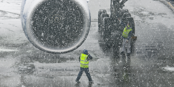 Two workers wearing high visibility jackets and winter gear work on a snowy tarmac next to a large aircraft.