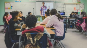 A teacher stands in a classroom addressing a dozen teenagers seated at desks with backpacks on the chairs. Photo by Kenny Eliason on Unsplash.