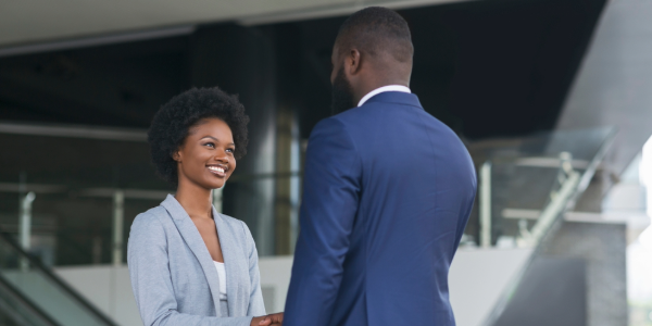 A young woman in a blue blazer smiles while shaking the hand of a man in a blue suit. They are standing in a modern office lobby.