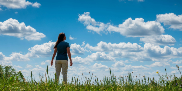 A woman wearing a blue t-shirt and tan pants stands in an open field and looks up at the blue sky.