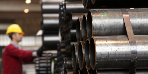 Steel pipes are stacked in the foreground while a worker in a red sweatshirt with a yellow hard hat is visible in the background.