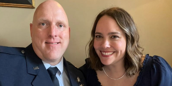 A man wearing a U.S. Air Force uniform smiles with his wife, who wears a blue top with a ruffled collar.