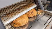 Two loaves of bread being sliced in an industrial slicing machine.