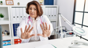 Brunette female in white lab coat raises hands to protect herself.