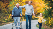 A middle-aged man walks with his father and son down a road with fall foliage in the background.