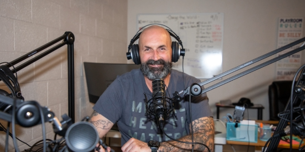 George Nolan sits at a desk wearing headphones with a recording microphone near his mouth. 