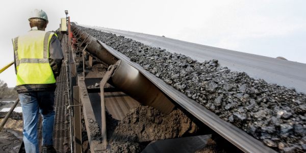 Worker wearing reflective vest and helmet walks alongside a coal-carrying conveyor belt.