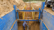 Worker at bottom of deep excavation guarded by a steel trench box to prevent trench collapse.