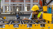 Female mechanical engineer in safety suit inspecting a machine in metal sheet production system.