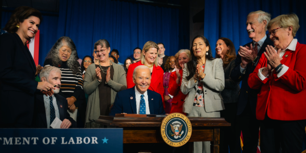 Surrounded by a dozen people applauding, President Biden looks up to smile while signing a proclamation at a desk bearing the presidential seal. 