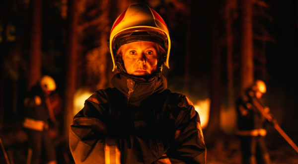 A female firefighter wearing her uniform and protective gear battling a fire in a smoky forest.