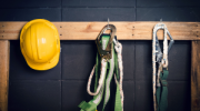 Yellow safety helmet and two safety harnesses hanging from pegs on a black wall. 