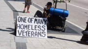 Older man in a baseball cap sits on a sidewalk under a small tree, next to a cart with belongings and has a large white cardboard sign that reads “one of many homeless veterans.” 