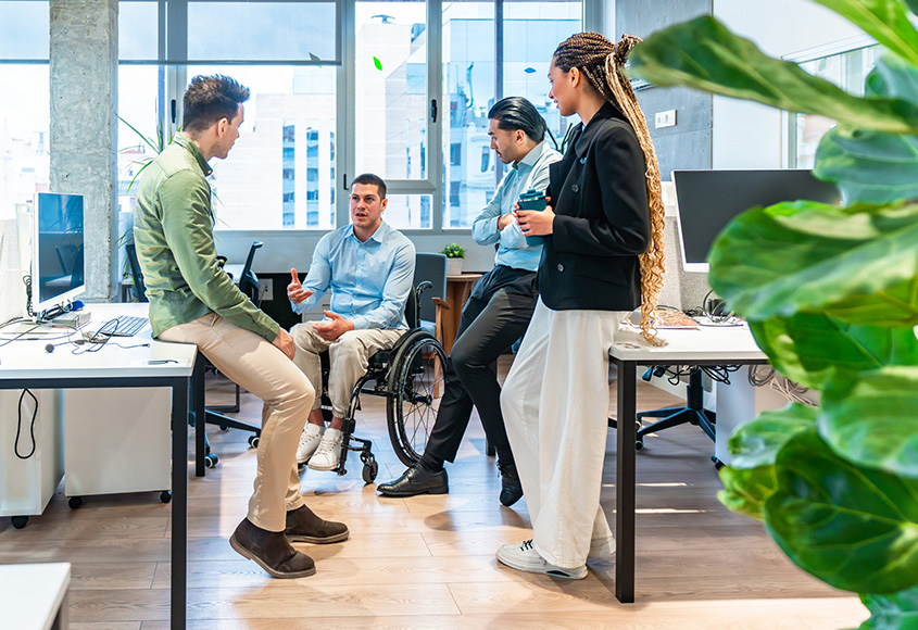 Several people, one in wheelchair, standing and sitting in office having a discussion.