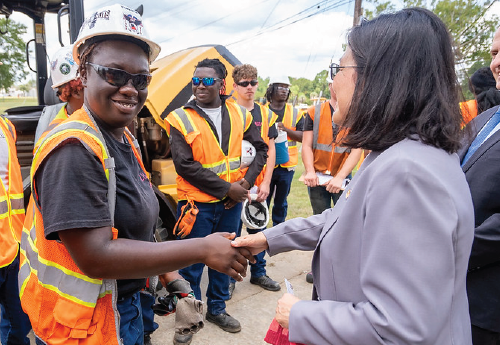 A woman in safety vest and hardhat smiles as she shakes Acting Secretary Su’s hand. More construction workers are gathered in the background. 