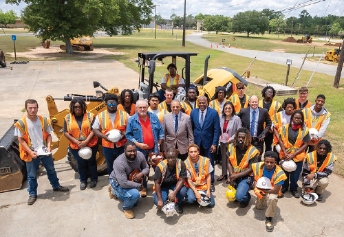 Two dozen people – some in suits, some in high-visibility vests and hard hats, pose for a photo in a park, gathered in front of an enormous track loader. 