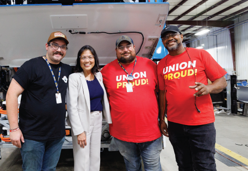 Acting Secretary Julie Su stands with three men – two wearing ‘union proud’ tee shirts – in front of a bus whose back panel is raised to expose its workings. 