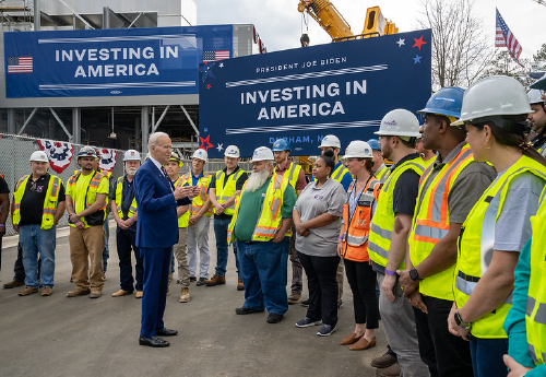 President Joe Biden addresses a dozen workers in high-visibility jackets and hard hats in front of construction equipment and signs reading Investing in America.