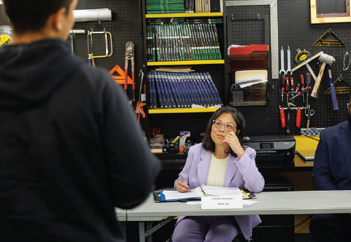 Seated at a table in a workshop with tools hung on the wall behind her, Acting Secretary Su listens to a worker speak. 