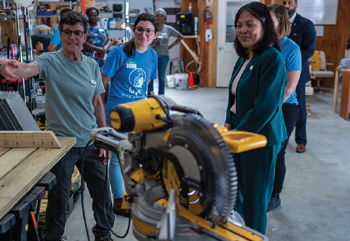 Acting Secretary Su smiles on a tour of a workshop. Two women gesture towards a work table beside a circular saw. 