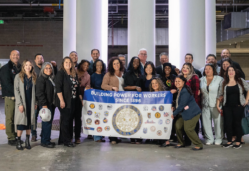 More than a dozen people pose for a photo in a building, holding a sign that says “building power for workers since 1896. 