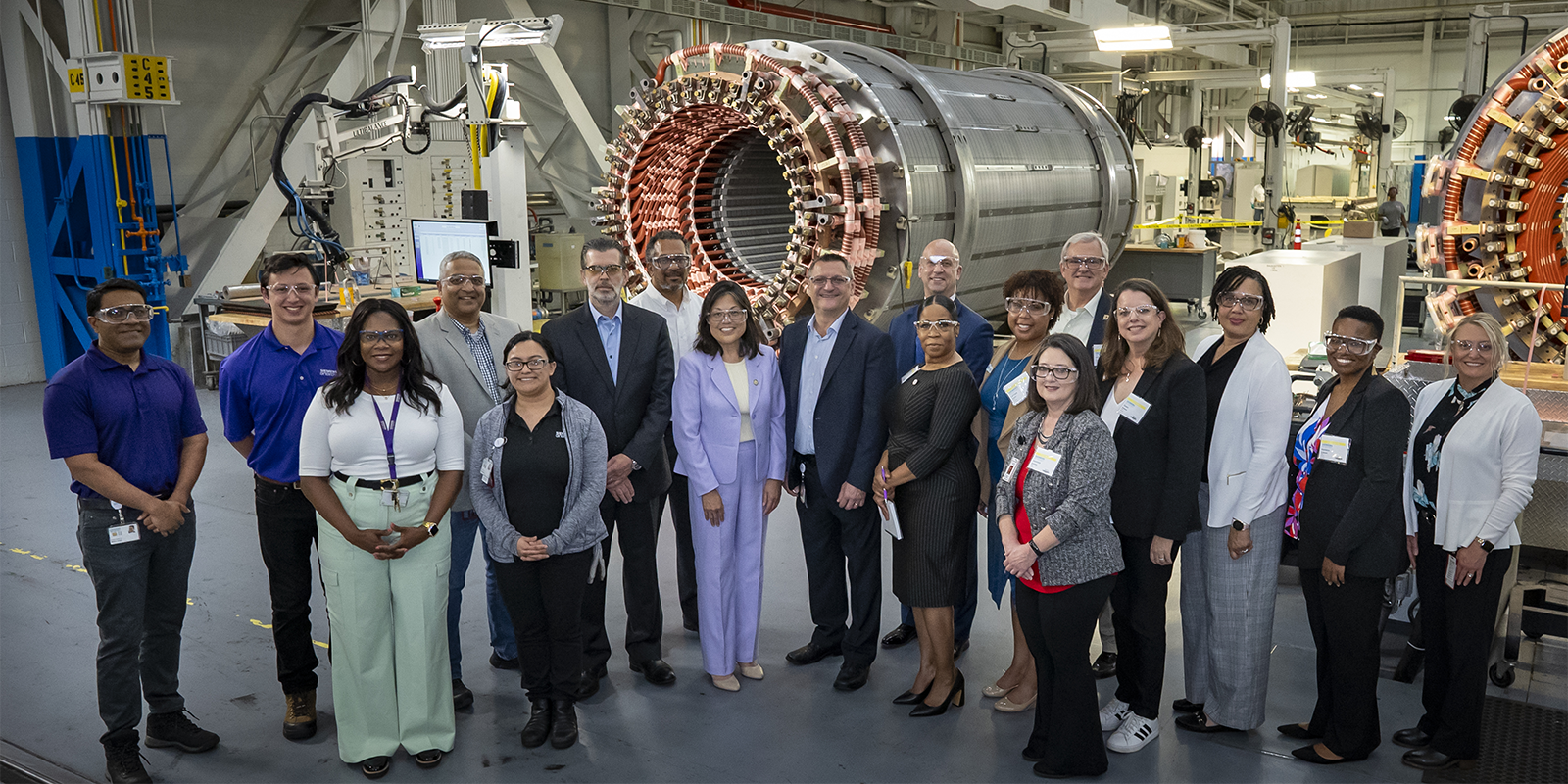 Acting Secretary Su poses with 17 men and women in professional clothing in a hangar where enormous high-tech metal cylinders are being constructed. 