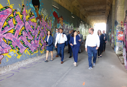 A dozen adults in professional clothing walk through an underpass whose walls are decorated with colorful graffiti. 