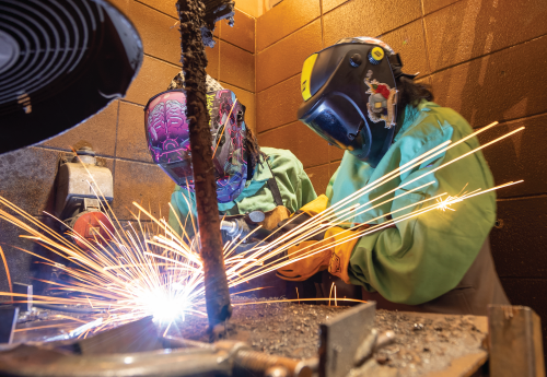 Sparks fly as two women in protective smocks, gloves and helmets are welding. 