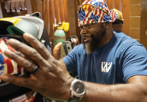 A man in a blue tee shirt and American flag cap fits a welding helmet on Acting Secretary Su. 