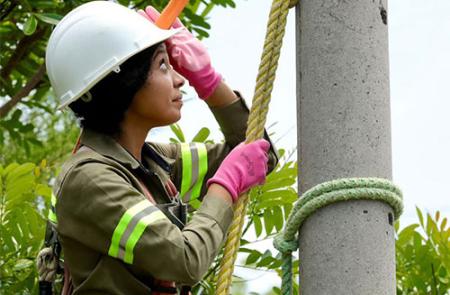 A woman in uniform and a hard hat uses a rope to anchor herself to a pole. Tree tops are in the background.